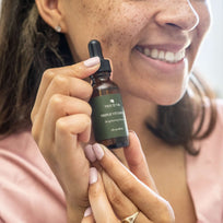 Brown skinned woman with freckles holding up the Vitamin C Serum while smiling