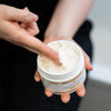 Closeup of opened Moisture Defense Shea Body Butter held by a woman showing the thick and rich texture of body butter on her fingers.